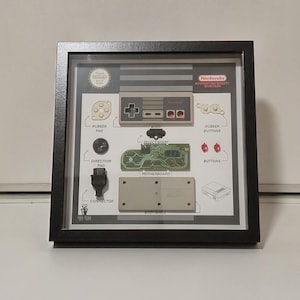 Puede incluir: Obra de arte enmarcada que muestra los componentes de un mando de Nintendo Entertainment System. La imagen incluye el cuerpo, la placa base, las almohadillas de goma, los botones y el conector, todos etiquetados. El logotipo de Nintendo es visible. El marco es negro.