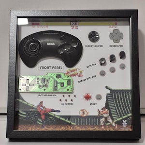 Puede incluir: Obra de arte enmarcada con un mando negro de Sega Genesis y componentes del videojuego Street Fighter II. Incluye una placa base, botones y un gráfico de una escena de lucha. El marco es negro.