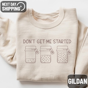 May include: Sand-colored sweatshirt with the phrase "DON'T GET ME STARTED" printed across the chest. The design features three jars with varying amounts of dots inside. The sweatshirt has a crew neck and long sleeves.