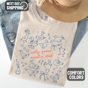 May include: Ivory-colored t-shirt with a blue goose illustration and the text "silly goose auntie club." The shirt is folded on top of blue jeans, with a tan hat and dried flowers in the background. The shirt is labeled "Comfort Colors."