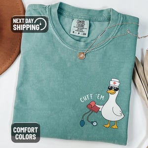 May include: Light green Comfort Colors t-shirt with a graphic of a duck wearing sunglasses and a nurse's hat, holding a blood pressure cuff. The text "CUFF 'EM" is printed above the duck. A gold necklace is draped on the shirt.