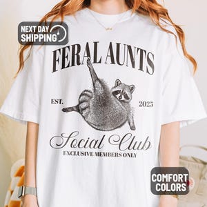 May include: White t-shirt with a graphic of a raccoon and the text "FERAL AUNTS Social Club". The design includes the year "2025" and the words "EXCLUSIVE MEMBERS ONLY". The shirt is labeled "COMFORT COLORS".