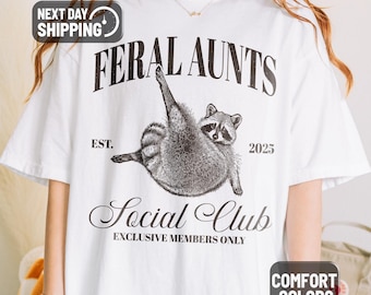May include: White t-shirt with a graphic of a raccoon and the text "FERAL AUNTS Social Club". The design includes the year "2025" and the words "EXCLUSIVE MEMBERS ONLY". The shirt is labeled "COMFORT COLORS".