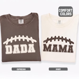 May include: Two folded t-shirts, one espresso and one ivory, each with a football design. The espresso shirt says "DADA" and the ivory shirt says "MAMA". The shirts have the words "COMFORT COLORS" printed on them.