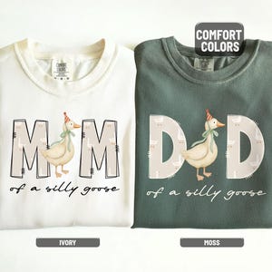 May include: Ivory and moss green crewneck sweatshirts. The ivory sweatshirt says "MOM of a silly goose" with a goose graphic. The moss green sweatshirt says "DAD of a silly goose" with a goose graphic. Both have the words "COMFORT COLORS" on them.