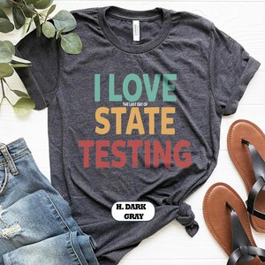 May include: Dark gray t-shirt with the text "I LOVE STATE TESTING" in teal, orange, and red. The shirt is paired with blue jeans and brown sandals. The text "H. DARK GRAY" is also visible.