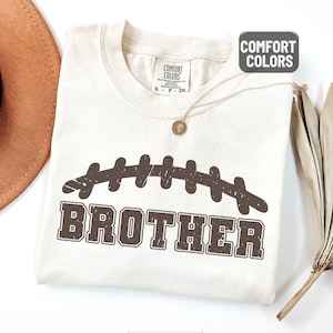 Puede incluir: Camiseta de color marfil con un dise&ntilde;o de bal&oacute;n de f&uacute;tbol marr&oacute;n y la palabra "BROTHER". La camiseta tiene cuello redondo y mangas cortas. La marca Comfort Colors es visible en la etiqueta.