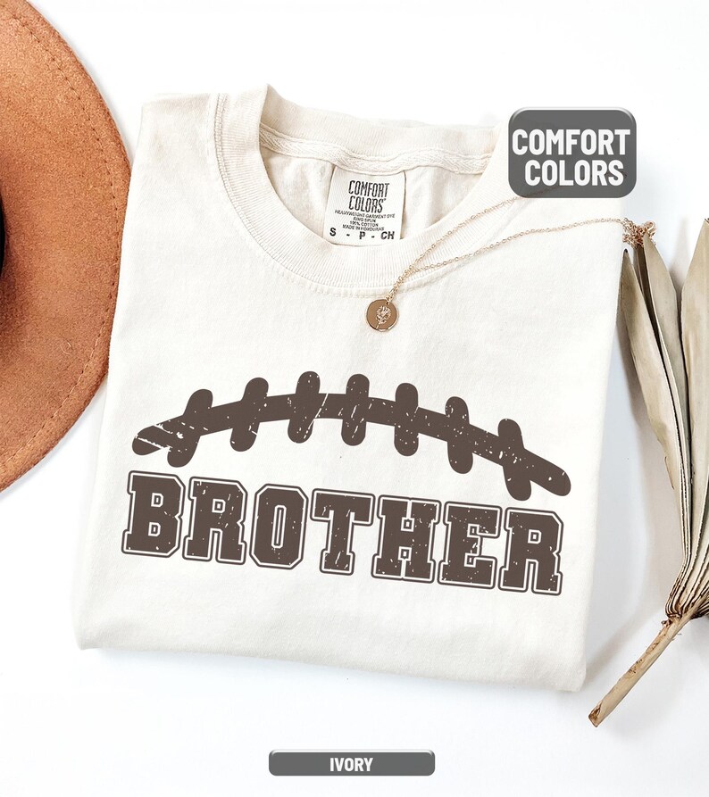 Puede incluir: Camiseta de color marfil con un dise&ntilde;o de bal&oacute;n de f&uacute;tbol marr&oacute;n y la palabra "BROTHER". La camiseta tiene cuello redondo y mangas cortas. La marca Comfort Colors es visible en la etiqueta.