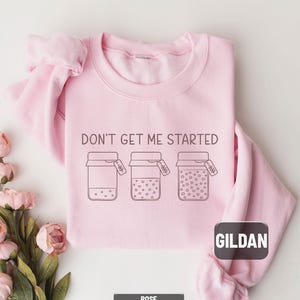 May include: A light pink Gildan sweatshirt with the phrase "DON'T GET ME STARTED" printed in black. Below the text are three jar illustrations with varying dot patterns. The sweatshirt is rose-colored.