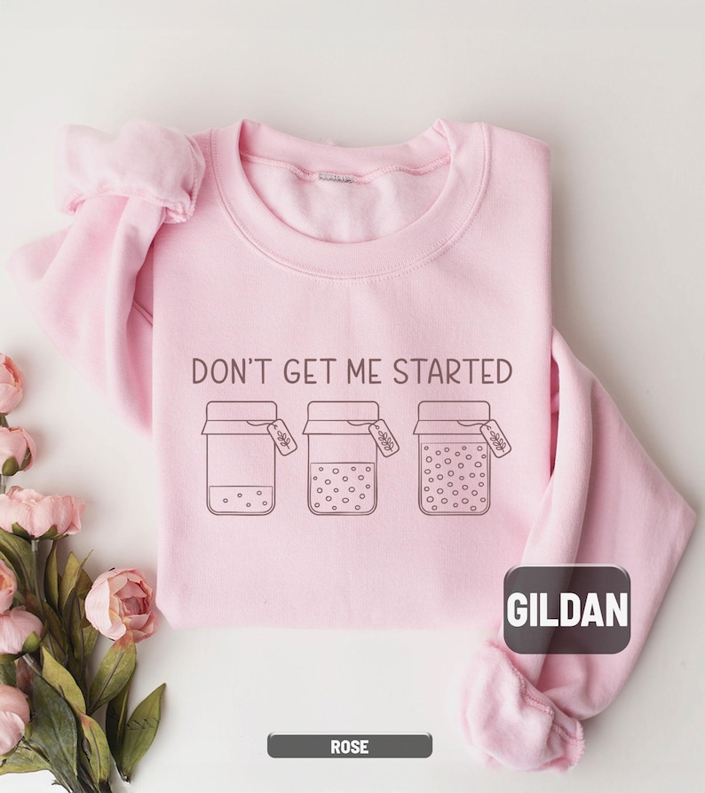 May include: A light pink Gildan sweatshirt with the phrase "DON'T GET ME STARTED" printed in black. Below the text are three jar illustrations with varying dot patterns. The sweatshirt is rose-colored.