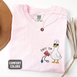 May include: A light pink Comfort Colors t-shirt with a cartoon duck wearing sunglasses and a nurse's hat, holding a blood pressure cuff. The text "CUFF 'EM" is above the duck. The shirt is labeled "Blossom".