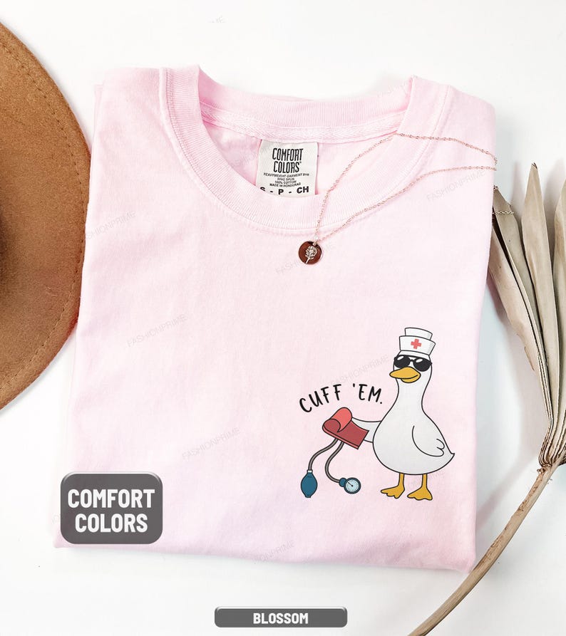 May include: A light pink Comfort Colors t-shirt with a cartoon duck wearing sunglasses and a nurse's hat, holding a blood pressure cuff. The text "CUFF 'EM" is above the duck. The shirt is labeled "Blossom".