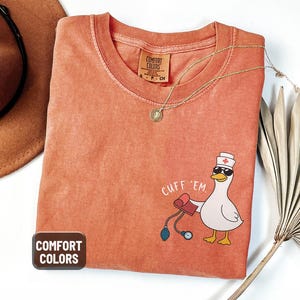May include: A coral-colored Comfort Colors t-shirt featuring a cartoon duck wearing sunglasses and a nurse's hat, holding a blood pressure cuff. The text "CUFF 'EM" is above the duck. A gold necklace and a brown hat are also visible.