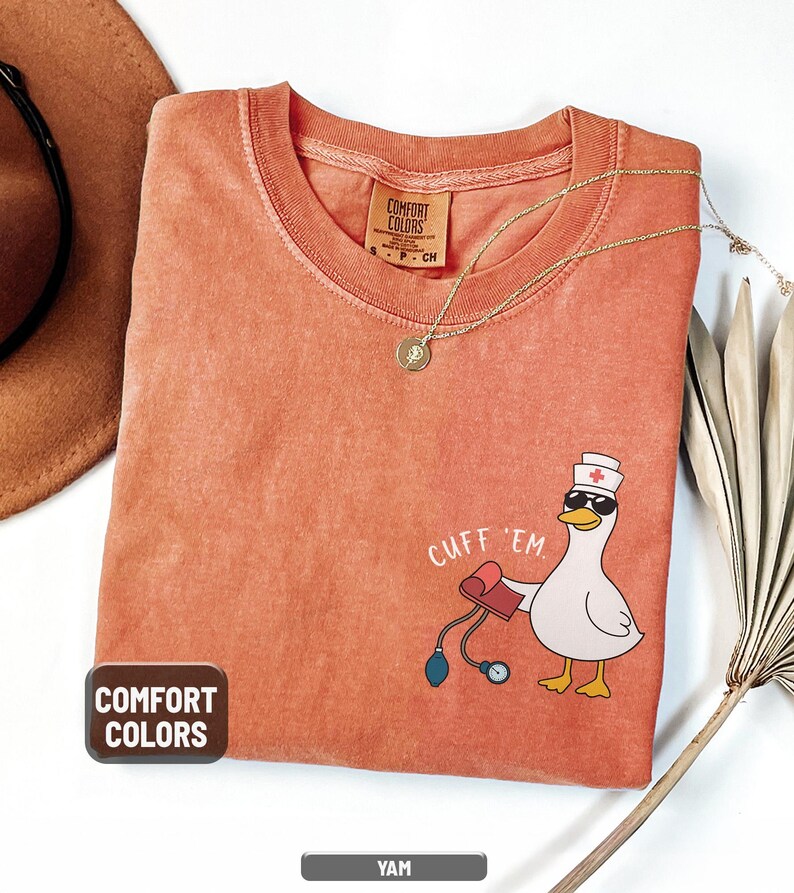 May include: A coral-colored Comfort Colors t-shirt featuring a cartoon duck wearing sunglasses and a nurse's hat, holding a blood pressure cuff. The text "CUFF 'EM" is above the duck. A gold necklace and a brown hat are also visible.