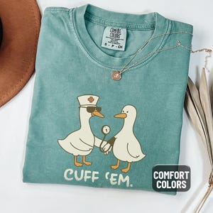 May include: Light green Comfort Colors t-shirt featuring a cartoon design of two ducks. One duck is wearing a nurse's hat and sunglasses, holding a blood pressure cuff. The text "CUFF 'EM" is printed below the ducks.
