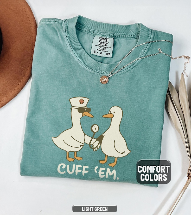 May include: Light green Comfort Colors t-shirt featuring a cartoon design of two ducks. One duck is wearing a nurse's hat and sunglasses, holding a blood pressure cuff. The text "CUFF 'EM" is printed below the ducks.