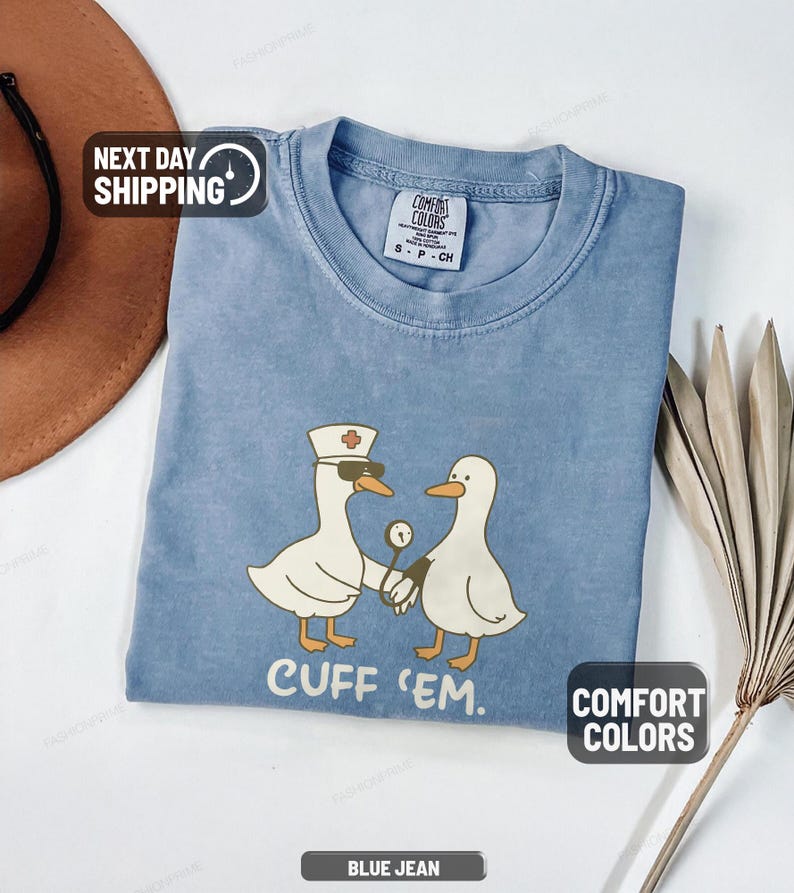 May include: Blue jean Comfort Colors t-shirt featuring a cartoon of two white geese. One goose wears a nurse's hat and sunglasses, holding a medical instrument. The text "CUFF 'EM" is below the geese.