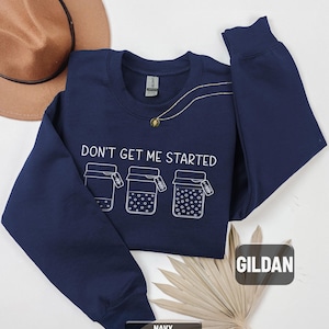 May include: Navy blue sweatshirt with the text "DON'T GET ME STARTED" and three illustrated jars. A gold necklace and a brown hat are also visible. The sweatshirt is made of a soft material.