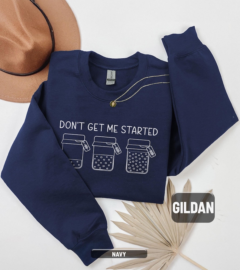 May include: Navy blue sweatshirt with the text "DON'T GET ME STARTED" and three illustrated jars. A gold necklace and a brown hat are also visible. The sweatshirt is made of a soft material.