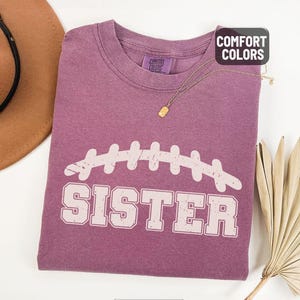 Puede incluir: Camiseta de color bayas con un dise&ntilde;o de f&uacute;tbol americano blanco y la palabra "SISTER" impresa debajo. La camiseta tiene cuello redondo y mangas cortas. Un collar dorado est&aacute; drapeado sobre la camiseta. La camiseta est&aacute; hecha de un material suave y c&oacute;modo.