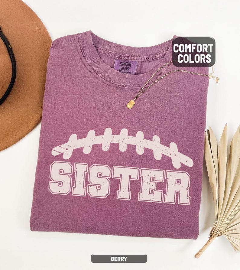 Puede incluir: Camiseta de color bayas con un dise&ntilde;o de f&uacute;tbol americano blanco y la palabra "SISTER" impresa debajo. La camiseta tiene cuello redondo y mangas cortas. Un collar dorado est&aacute; drapeado sobre la camiseta. La camiseta est&aacute; hecha de un material suave y c&oacute;modo.