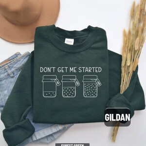May include: Forest green sweatshirt with the text "DON'T GET ME STARTED" and three jars with varying amounts of dots. The sweatshirt is a Gildan brand. A brown hat and blue jeans are in the background.