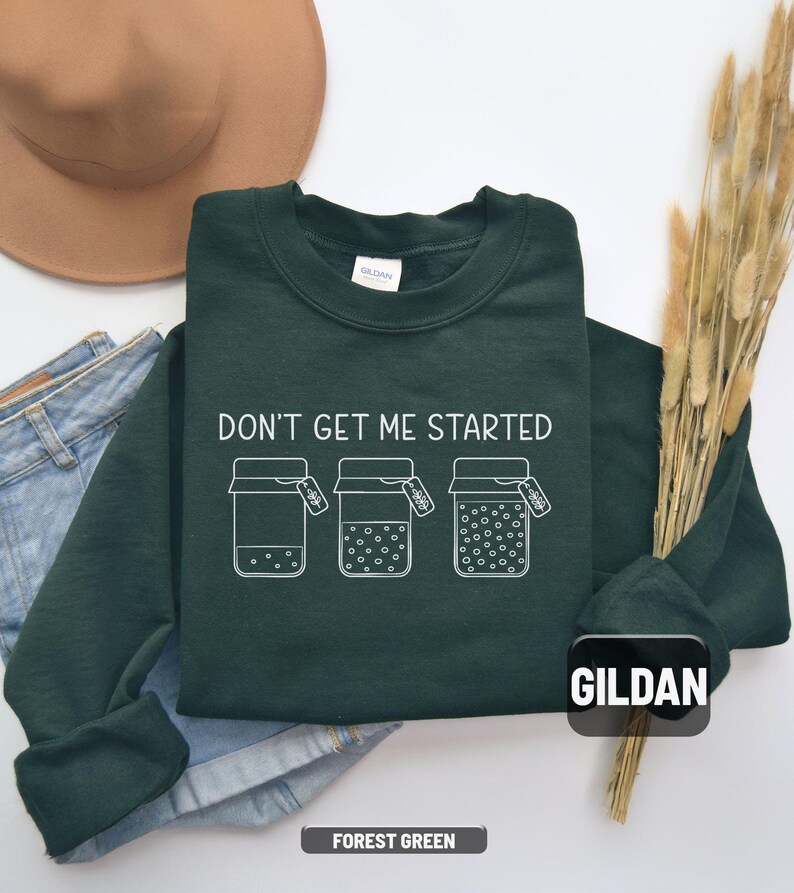 May include: Forest green sweatshirt with the text "DON'T GET ME STARTED" and three jars with varying amounts of dots. The sweatshirt is a Gildan brand. A brown hat and blue jeans are in the background.