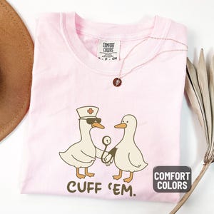 May include: A light pink t-shirt with a graphic of two cartoon ducks, one wearing a nurse's hat and sunglasses, holding a medical instrument. The text "CUFF 'EM" is below the ducks. The shirt has the "Comfort Colors" label.
