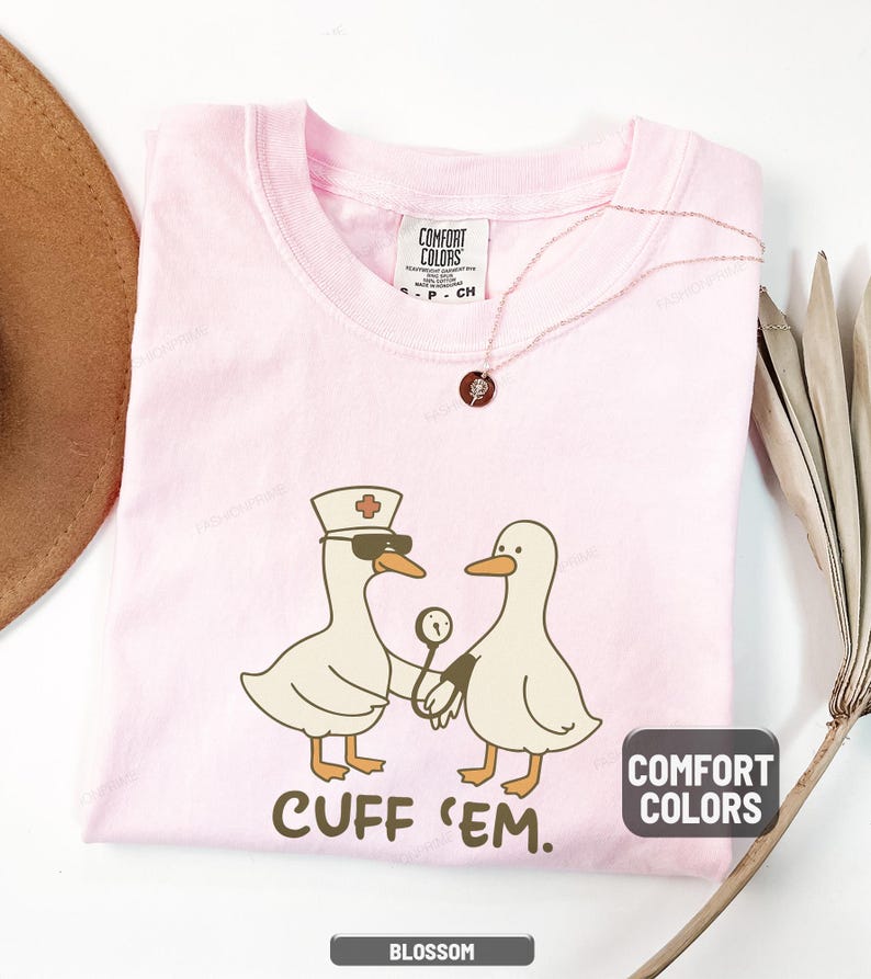 May include: A light pink t-shirt with a graphic of two cartoon ducks, one wearing a nurse's hat and sunglasses, holding a medical instrument. The text "CUFF 'EM" is below the ducks. The shirt has the "Comfort Colors" label.