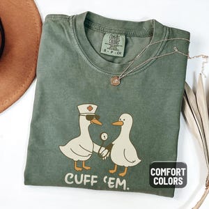 May include: Moss green Comfort Colors t-shirt with a graphic of two cartoon ducks, one wearing a nurse's hat and sunglasses, holding a stethoscope. The text "CUFF 'EM" is below the ducks. A gold necklace is draped on the shirt.