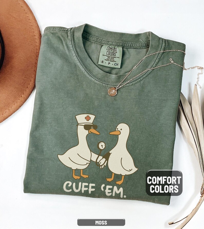 May include: Moss green Comfort Colors t-shirt with a graphic of two cartoon ducks, one wearing a nurse's hat and sunglasses, holding a stethoscope. The text "CUFF 'EM" is below the ducks. A gold necklace is draped on the shirt.
