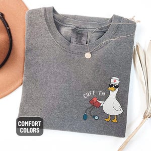 May include: A gray Comfort Colors t-shirt with a cartoon duck wearing sunglasses and a nurse's hat, holding a blood pressure cuff. The text "CUFF 'EM" is above the duck. A gold necklace and a brown hat are also visible.