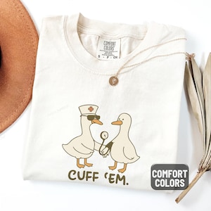 May include: Ivory-colored t-shirt with a humorous graphic of two cartoon ducks, one wearing a nurse's hat and sunglasses, holding a stethoscope. The text "CUFF 'EM" is below the ducks. The shirt has a Comfort Colors label.