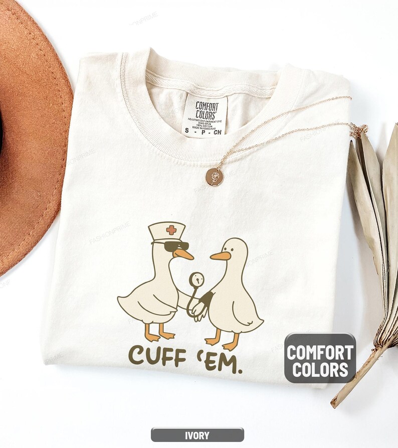 May include: Ivory-colored t-shirt with a humorous graphic of two cartoon ducks, one wearing a nurse's hat and sunglasses, holding a stethoscope. The text "CUFF 'EM" is below the ducks. The shirt has a Comfort Colors label.