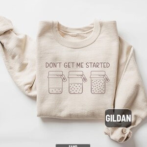 May include: Sand-colored sweatshirt with the phrase "DON'T GET ME STARTED" printed across the chest. The design features three jars with varying amounts of dots inside. The brand name "GILDAN" is visible on the lower right.