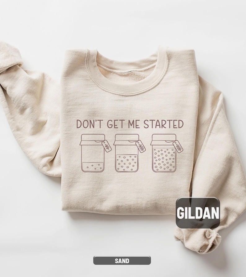 May include: Sand-colored sweatshirt with the phrase "DON'T GET ME STARTED" printed across the chest. The design features three jars with varying amounts of dots inside. The brand name "GILDAN" is visible on the lower right.