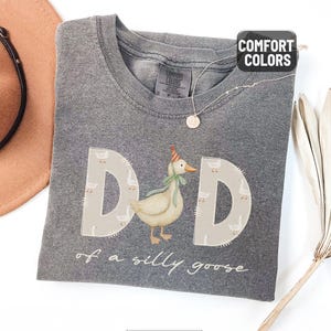May include: A gray Comfort Colors t-shirt with the word "DAD" in large letters, featuring a goose wearing a party hat and a green bow tie. The text "of a silly goose" is below. A brown hat and necklace are visible.