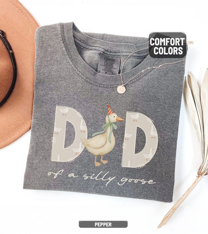 May include: A gray Comfort Colors t-shirt with the word "DAD" in large letters, featuring a goose wearing a party hat and a green bow tie. The text "of a silly goose" is below. A brown hat and necklace are visible.