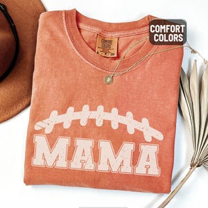 Puede incluir: Una camiseta de color coral con un gr&aacute;fico de f&uacute;tbol desgastado y la palabra "MAMA" en negrita. La camiseta tiene cuello redondo y mangas cortas. La etiqueta dice "Comfort Colors". Tambi&eacute;n se ven un collar dorado y un sombrero marr&oacute;n.