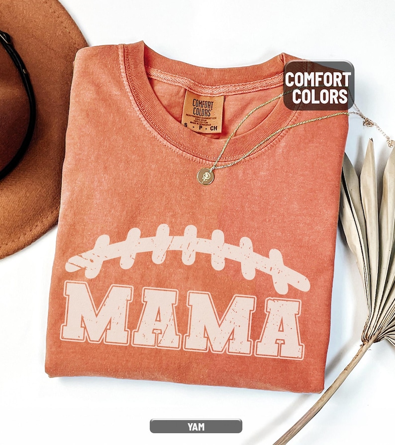 Puede incluir: Una camiseta de color coral con un gr&aacute;fico de f&uacute;tbol desgastado y la palabra "MAMA" en negrita. La camiseta tiene cuello redondo y mangas cortas. La etiqueta dice "Comfort Colors". Tambi&eacute;n se ven un collar dorado y un sombrero marr&oacute;n.