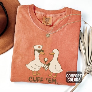 May include: A yam-colored Comfort Colors t-shirt featuring a cartoon duck wearing sunglasses and a nurse's hat, holding a medical instrument, with another duck. The text "CUFF 'EM" is below the ducks. A gold necklace and a brown hat are also visible.