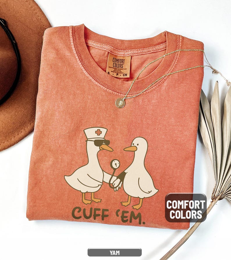 May include: A yam-colored Comfort Colors t-shirt featuring a cartoon duck wearing sunglasses and a nurse's hat, holding a medical instrument, with another duck. The text "CUFF 'EM" is below the ducks. A gold necklace and a brown hat are also visible.