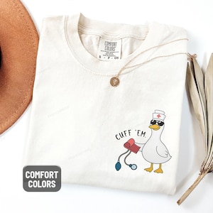 May include: Ivory-colored t-shirt with a cartoon goose wearing sunglasses and a nurse's hat, holding a blood pressure cuff. The text "CUFF 'EM" is printed above the goose. The shirt has a Comfort Colors tag.