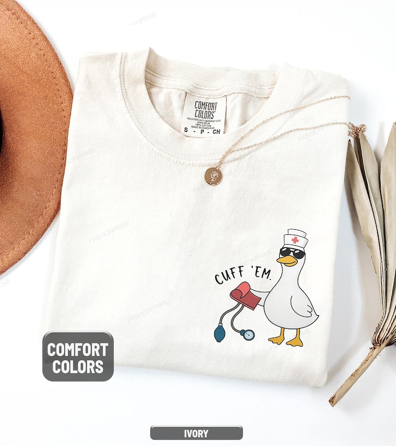 May include: Ivory-colored t-shirt with a cartoon goose wearing sunglasses and a nurse's hat, holding a blood pressure cuff. The text "CUFF 'EM" is printed above the goose. The shirt has a Comfort Colors tag.