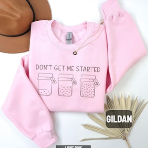May include: Light pink sweatshirt with the phrase "DON'T GET ME STARTED" and three jar illustrations. The sweatshirt is a crew neck style and features the brand name "GILDAN". The bottom of the sweatshirt is labeled "LIGHT PINK".