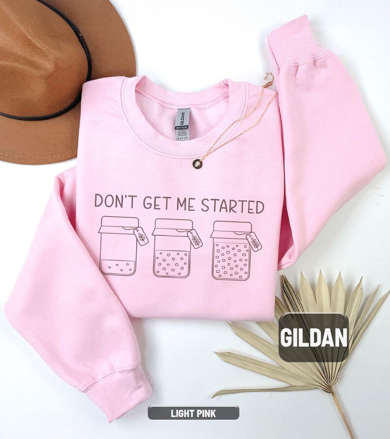 May include: Light pink sweatshirt with the phrase "DON'T GET ME STARTED" and three jar illustrations. The sweatshirt is a crew neck style and features the brand name "GILDAN". The bottom of the sweatshirt is labeled "LIGHT PINK".