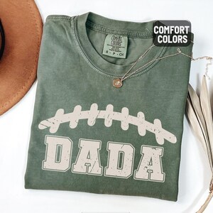 Puede incluir: Una camiseta verde musgo con un gr&aacute;fico de f&uacute;tbol desgastado y la palabra "DADA" en una fuente vintage. La camiseta tiene cuello redondo y mangas cortas. Un collar dorado con un peque&ntilde;o colgante est&aacute; sobre la camiseta.