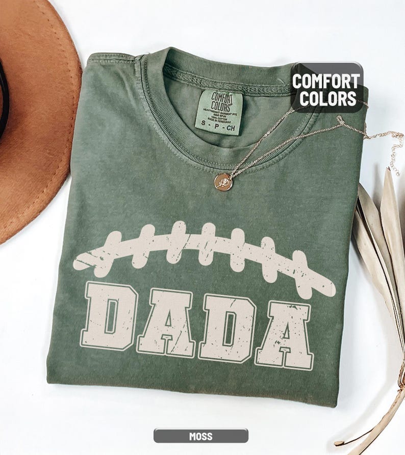 Puede incluir: Una camiseta verde musgo con un gr&aacute;fico de f&uacute;tbol desgastado y la palabra "DADA" en una fuente vintage. La camiseta tiene cuello redondo y mangas cortas. Un collar dorado con un peque&ntilde;o colgante est&aacute; sobre la camiseta.