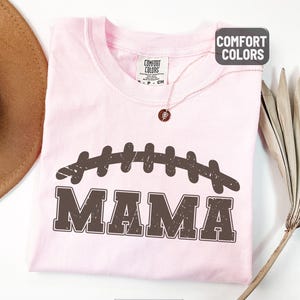 Puede incluir: Camiseta rosa claro con un dise&ntilde;o de bal&oacute;n de f&uacute;tbol marr&oacute;n desgastado y la palabra "MAMA" impresa debajo. La camiseta tiene cuello redondo y mangas cortas. La palabra "BLOSSOM" est&aacute; impresa en la parte inferior de la imagen.