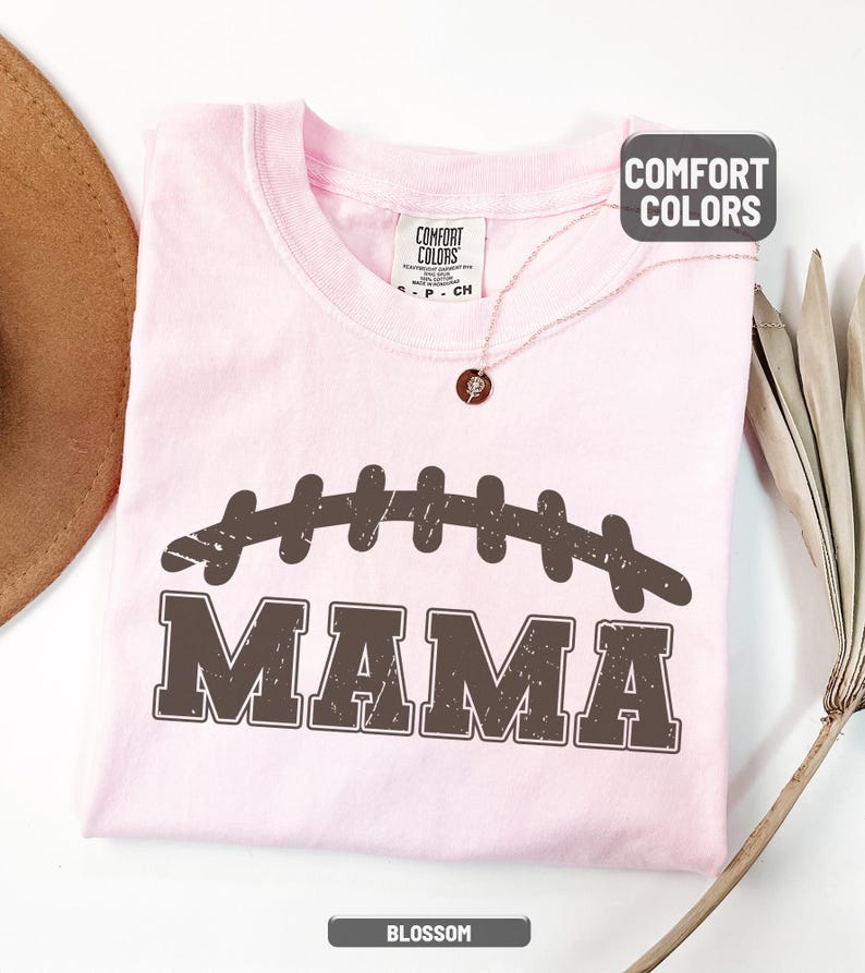 Puede incluir: Camiseta rosa claro con un dise&ntilde;o de bal&oacute;n de f&uacute;tbol marr&oacute;n desgastado y la palabra "MAMA" impresa debajo. La camiseta tiene cuello redondo y mangas cortas. La palabra "BLOSSOM" est&aacute; impresa en la parte inferior de la imagen.